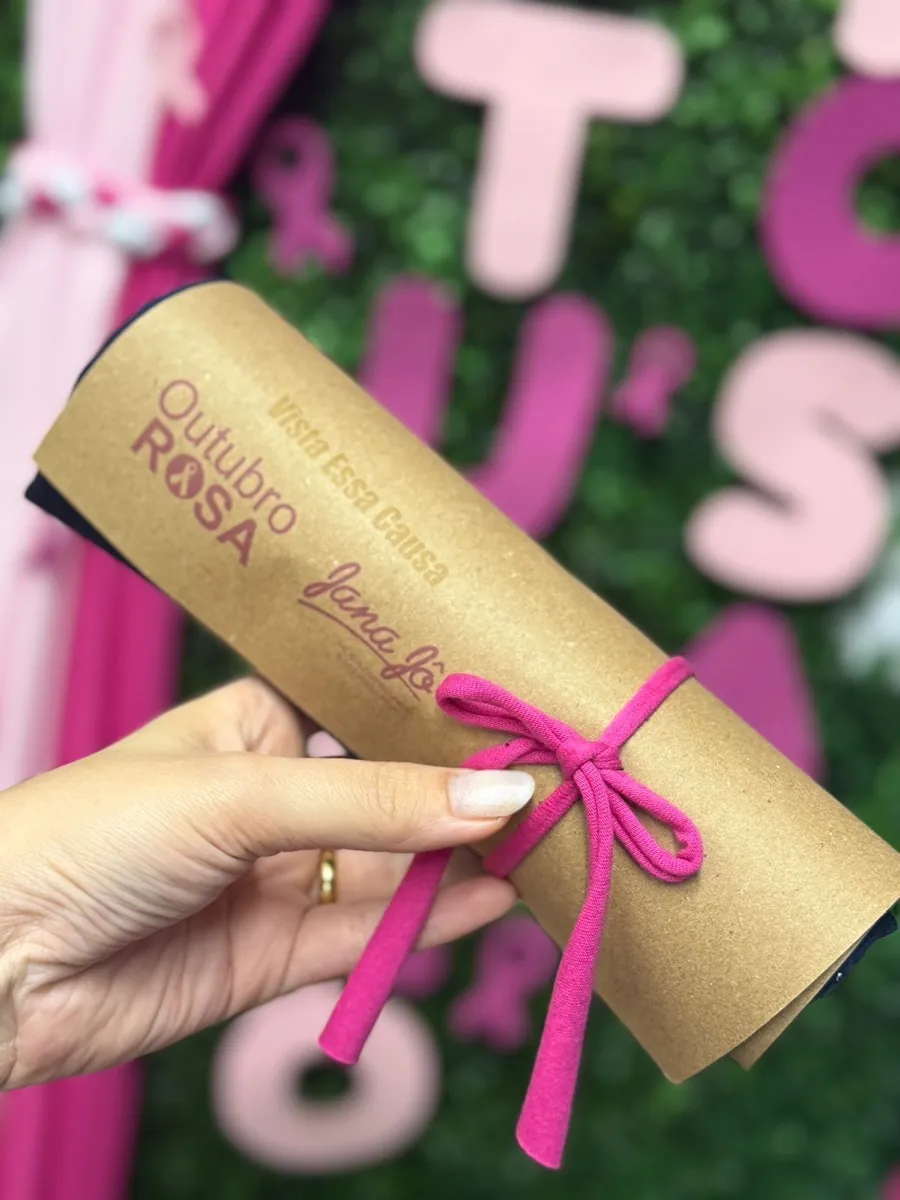 Camiseta enrolada no papel kraft da campanha, amarrada com um laço de malha rosa e impressa com Outubro Rosa e Vista Essa Causa